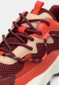 Red and beige athletic shoe with mesh and leather materials, featuring intricate red lacing, a padded collar, and a textured sole.