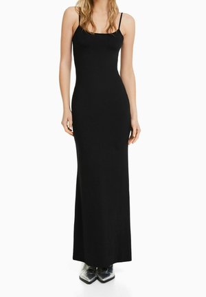 Woman wearing a floor-length black sleeveless dress with thin straps and black pointed-toe boots on a plain white background.