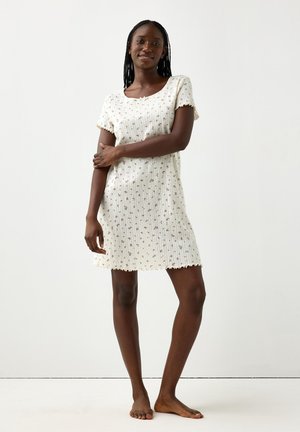 Femme debout pieds nus portant une robe crème à manches courtes, longueur genou, avec de petits motifs floraux sur fond blanc.