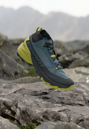 Trail running cipela plave i žute boje s potporom za gležanj i vezicama, lebdi iznad kamenitog planinskog terena.