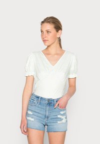 Blusa de mangas curtas azul claro com decote em V e bolinhas amarelas. Estilizada com calções de ganga desgastados de cintura alta.