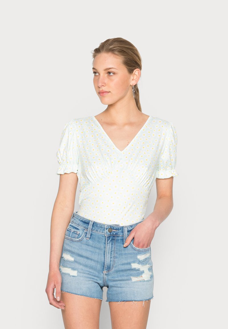 Blusa de mangas curtas azul claro com decote em V e bolinhas amarelas. Estilizada com calções de ganga desgastados de cintura alta.