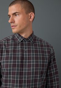 Dark plaid shirt featuring tones of maroon, navy, and white, with a button-down collar and front button closure, made from a textured fabric.