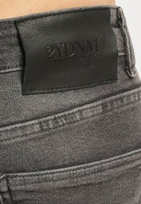 Grå denimjeans med en svart lærlapp med preget tekst. Merkbare detaljer inkluderer sømmer og lommeomriss.