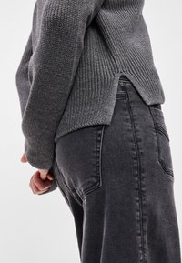 Sweater cinza canelado com gola alta e fenda lateral, combinado com jeans cinza escuro, destacando texturas e detalhes ajustados.