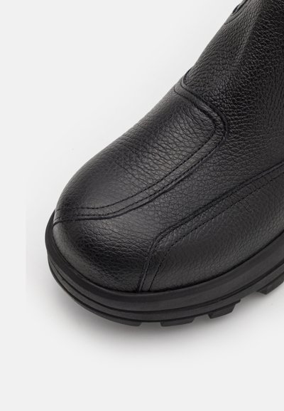 Bota de cuero negro con un acabado texturizado, punta redondeada y suela de goma negra gruesa con un patrón de banda de rodadura. Costura visible en la parte superior.