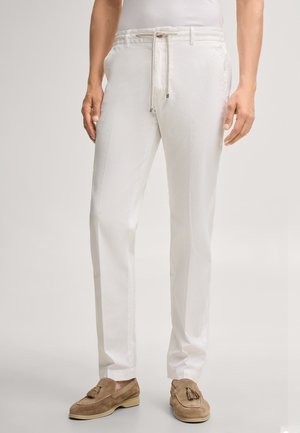 Mężczyzna ubrany w białe spodnie ze ściągaczem i beżowe zamszowe mokasyny z frędzlami, stojący na jednolitym jasnym tle.