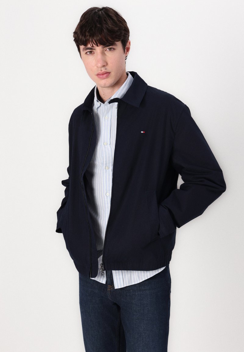 Navyblaues Jackett mit Kragen, Frontreißverschluss, zwei Taschen und kleinem Logo. Getragen über einem hellblauen gestreiften Hemd und Jeans.