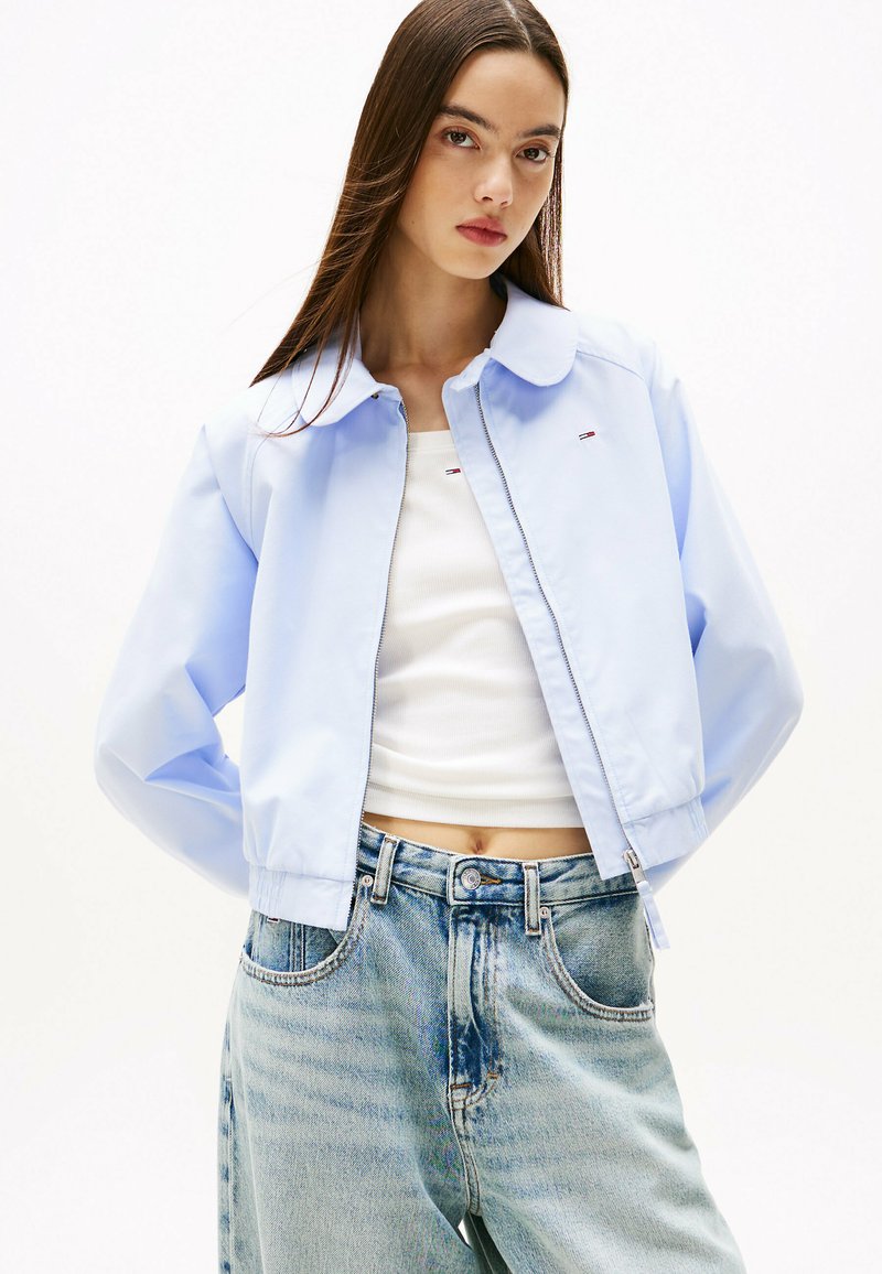 Giacca cropped azzurra con colletto classico e chiusura con zip, caratterizzata da un sottile dettaglio del logo, abbinata a una canotta bianca e jeans in denim strappati.