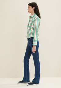 Gestreifte Button-Down-Bluse in Grün, Weiß und Rot mit einer Brusttasche. Kombiniert mit dunkelblauen Schlagjeans und marineblauen Sneakers.