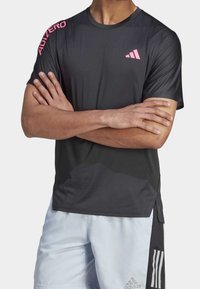 Svart träningströja med korta ärmar, rund hals och rosa Adidas-logotyper. Tillverkad av lätt och andningsbart material.