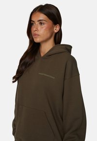 Oliwkowa bluza z kapturem z przednią kieszenią, wykonana z miękkiej tkaniny. Posiada logo w jaśniejszym odcieniu na klatce piersiowej. Luźny, swobodny krój.