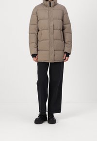 Personne portant une veste d'hiver matelassée taupe avec un col haut, un pantalon noir à jambes larges, et des chaussures noires massives à lacets, debout sur un fond blanc.