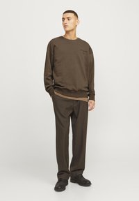 Sudadera marrón con cuello redondo y hombros caídos, combinada con pantalones marrones holgados. Unos zapatos negros completan el conjunto.