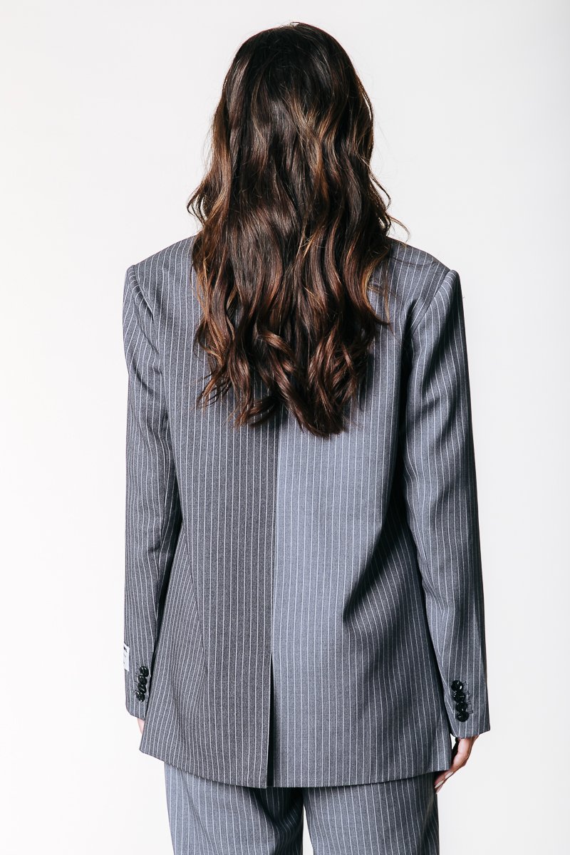 Chaqueta de traje a rayas grises con un diseño estructurado, una abertura trasera y acentos de botón negro en los puños. Tela texturizada visible.
