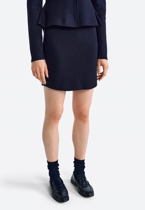 Vrouw draagt een marineblauwe rok, bijpassende top met lange mouwen, zwarte sokken en zwarte glanzende loafers, staand tegen een effen witte achtergrond.
