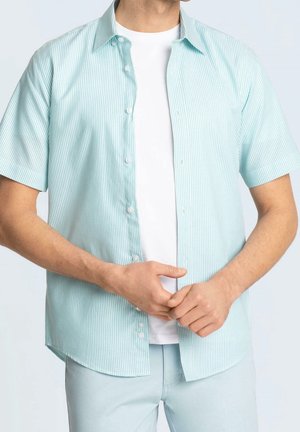 Mann trägt hellblau-weiß gestreiftes kurzärmeliges Hemd über einem schlichten weißen T-Shirt, die Hände vor dem Körper verschränkt.
