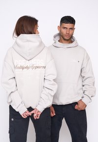 Haut de sweatshirt gris clair avec une grande poche avant, des poignets côtelés et une capuche avec cordon de serrage. Présente un texte graphique au dos et une marque discrète à l'avant.