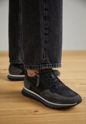 Sorte og grå ruskindssneakers med snørebånd og sidelås, båret med mørkegrå denimjeans på et lyst trægulv.