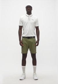 Άνδρας στέκεται με το πρόσωπο προς τα εμπρός φορώντας λευκό καπέλο, λευκό πόλο μπλουζάκι, σορτς ελιάς, μαύρη ζώνη, λευκές κάλτσες και λευκά παπούτσια.