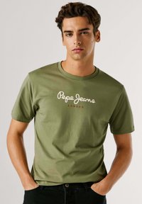 Alyvuogių spalvos medvilninis T-shirt su apvaliu iškirpimu, trumpomis rankovėmis, su baltu "Pepe Jeans London" logotipu ir raudona akcentu. Lygus paviršius.