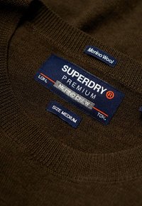 Pull col rond en laine mérinos brun foncé avec étiquettes de marque bleu et rouge, comprenant "SUPERDRY PREMIUM" et la taille "Medium". Tissu tricoté texturé.