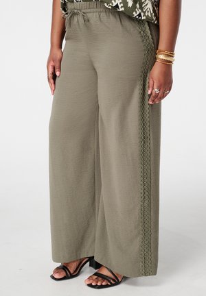 Femme portant un pantalon ample kaki avec un galon décoratif sur le côté, associé à des talons noirs à brides et des bracelets dorés au poignet.