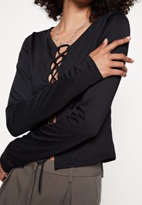 Top nero a maniche lunghe con scollatura a V profonda e dettagli in laccatura, tessuto morbido, maniche aderenti e orlo corto. Abbinato a pantaloni grigi.