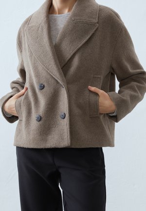 Personne portant un manteau en laine croisé couleur taupe avec des boutons noirs, les mains dans les poches, associé à un pantalon noir et une chemise grise en dessous.