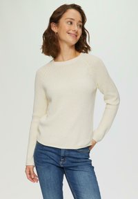 Pull en maille blanche avec texture côtelée, col rond et manches longues, associé à un jean bleu. Design décontracté et confortable.