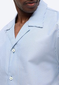 Άνδρας που φοράει ανοιχτό πουκάμισο με κάθετες ρίγες σε ανοιχτό μπλε και λευκό, με ανοιχτό γιακά, εμφανίζοντας μερικώς το πρόσωπο και τον λαιμό.