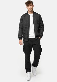 Veste bomber noire matelassée avec col côtelé, associée à un pantalon cargo noir avec poches et des chaussures de sport blanches.
