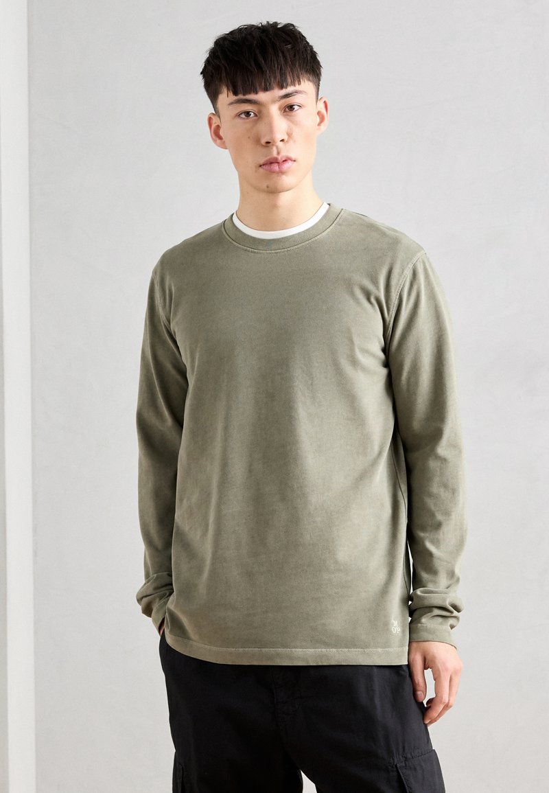 T-shirt à manches longues, vert olive, fabriqué en tissu doux, avec un col rond et un petit logo sur l'ourlet. Un pantalon noir complète le look.