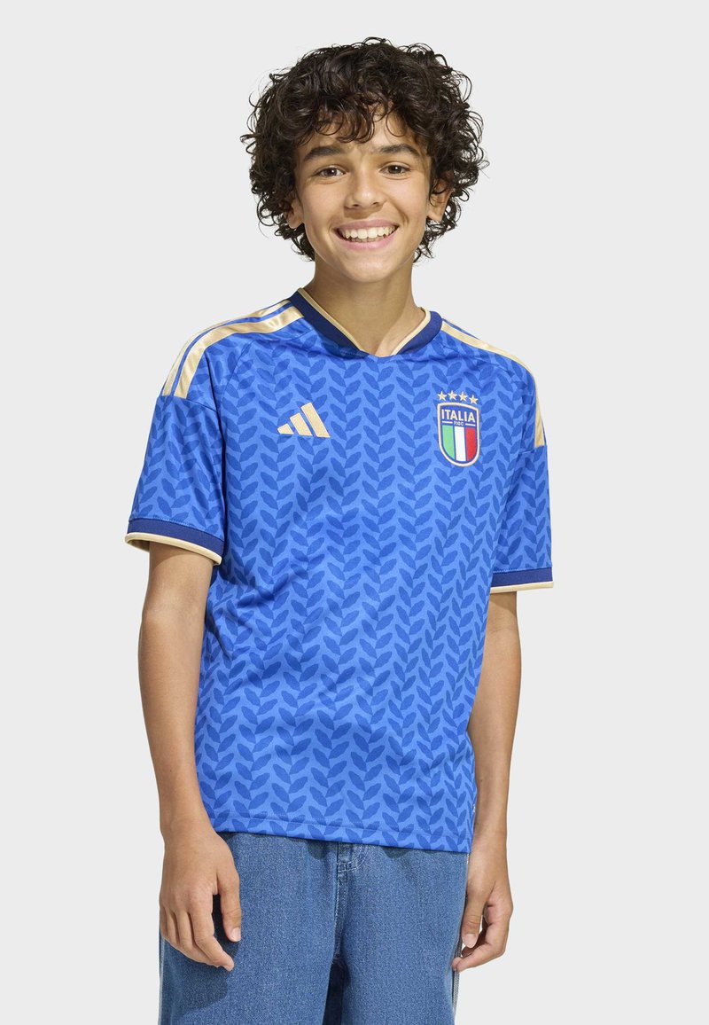 Blå kortärmad fotbollströja med ett texturerat lövmönster, gulddetaljer och en "Italia"-logotyp. Kombinerad med löst sittande jeansshorts.