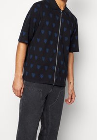 Polo shirt nera a maniche corte con chiusura a zip e un motivo a cuori nei colori blu e verde a forma di diamante. Realizzata in tessuto testurizzato.