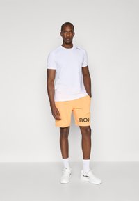 White athletic t-shirt, orange shorts with "BOR" printed in black, wearing white sneakers and white socks, standing on a plain background.