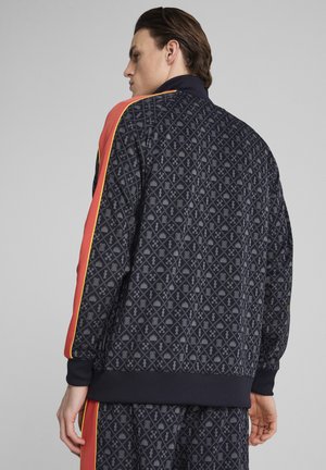 Trainingsjack met een geometrisch patroon in marineblauw, met oranje zijstrepen en gele accenten. Geribbelde manchetten en onderkant.