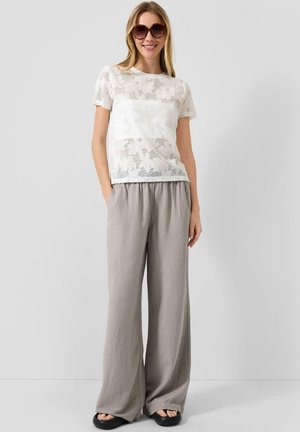 Kobieta nosząca duże okulary przeciwsłoneczne, biały kwiecisty koronkowy top z krótkim rękawem, luźne jasnoszare spodnie i czarne sandały, stojąca z rękami w kieszeniach.