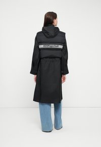 Zwarte gewatteerde jas met een capuchon, met een trekkoord in de taille. Grijs "H2OFagerholt" logo over de rug, gecombineerd met lichtblauwe jeans.