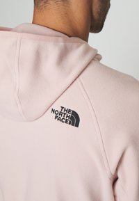 Light pink hoodie made of soft fabric, featuring a black embroidered logo on the back near the neckline and stitched detailing.