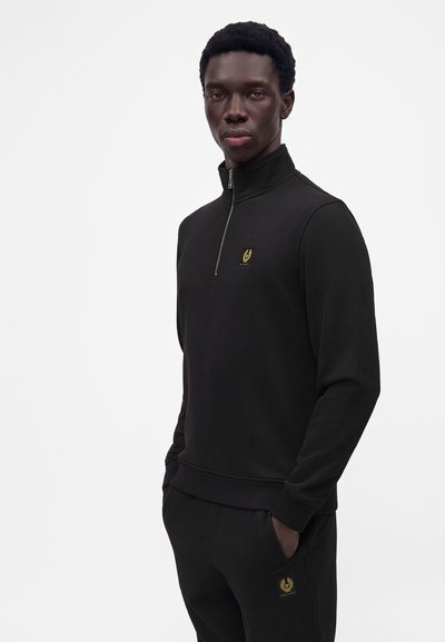 Jeune homme portant un sweat-shirt noir à demi-fermeture éclair et un pantalon assorti avec un logo de laurier doré, debout les mains dans les poches sur fond blanc.
