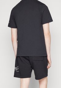 Osoba oblečená v černém krátkém rukávu tričku a černých šortkách s bílým nápisem, zobrazená zezadu na jednobarevném pozadí.