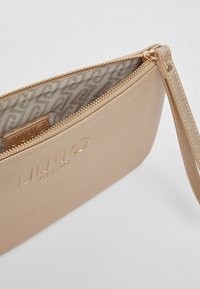 Pochette beige métallique en cuir texturé, fermeture éclair et bracelet pour poignet. Intérieur doublé avec un tissu à motif. Logo embossé sur le devant.