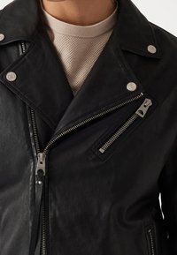 Schwarze Lederjacke mit silbernen Beschlägen, mit einem Reverskragen, zwei durchgehenden Reißverschlusstaschen und strukturiertem Stoff darunter.