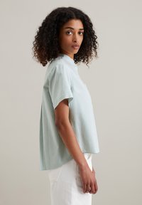 Camicetta azzurra chiara a maniche corte con righe bianche verticali, colletto e vestibilità rilassata. Il tessuto appare liscio con una leggera lucentezza.