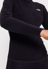 Maglietta nera in pile a maniche lunghe con zip a un quarto, dotata di un logo discreto sul petto e polsini aderenti. Texture liscia e design slim.
