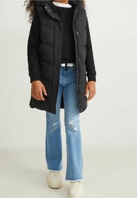 Dziecko w czarnej kurtce puchowej noszące czarną bluzkę z długim rękawem, jasnoniebieskie rozerwane dżinsy w stylu flare i białe tenisówki, stojące na tle jednolitego tła.