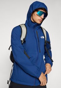 Μπλε αδιάβροχο μπουφάν με κουκούλα, τσέπη στο στήθος με φερμουάρ, ελαστικούς καρπούς και ρυθμιζόμενο κορδόνι. Φορεμένο πάνω από μια γκρίζα τσάντα πλάτης.