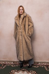 Donna con lunghi capelli rossi che indossa un lungo cappotto di pelliccia beige oversize, jeans blu e stivali neri a punta, in piedi su un tappeto decorato.