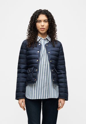 Kobieta o kręconych włosach, ubrana w granatową pikowaną kurtkę na niebiesko-białej koszuli w pionowe paski oraz ciemne dżinsy, stojąca na jednolitym tle.